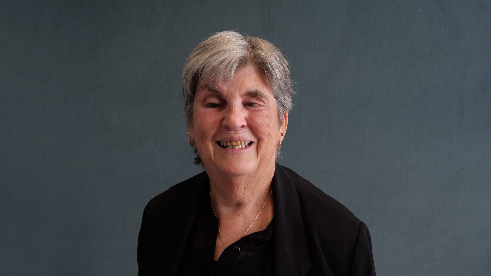 A woman with short grey hair, wearing a black shirt smiling for the camera with a dark grey internal background.