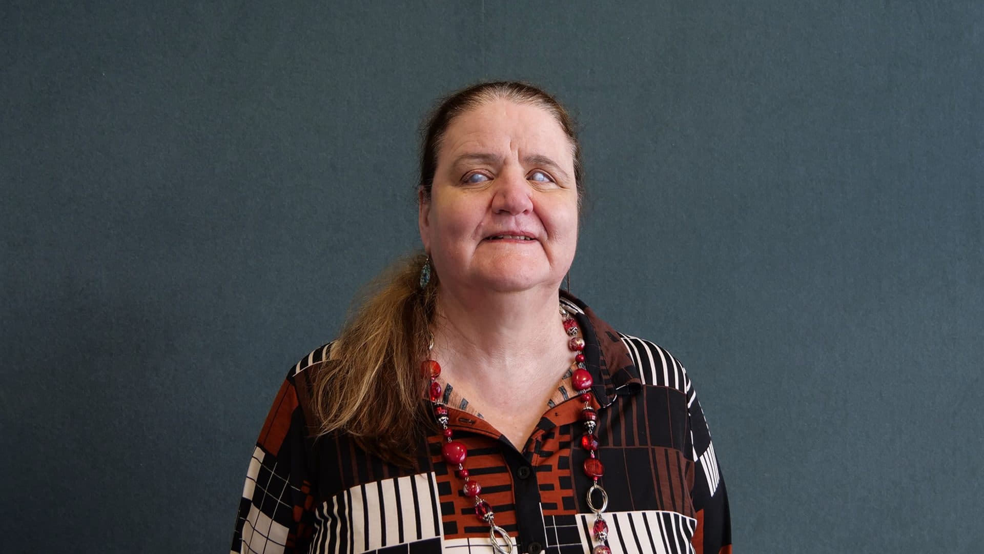 A woman with long red hair wearing glasses, a black, white, and brown patterned blouse smiling for the camera with a dark grey internal background.