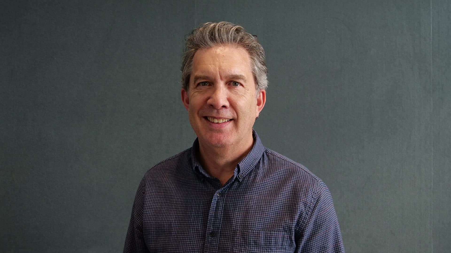 A man with greyhair, and a blue patterned shirt smiling for the camera with a dark grey internal background.