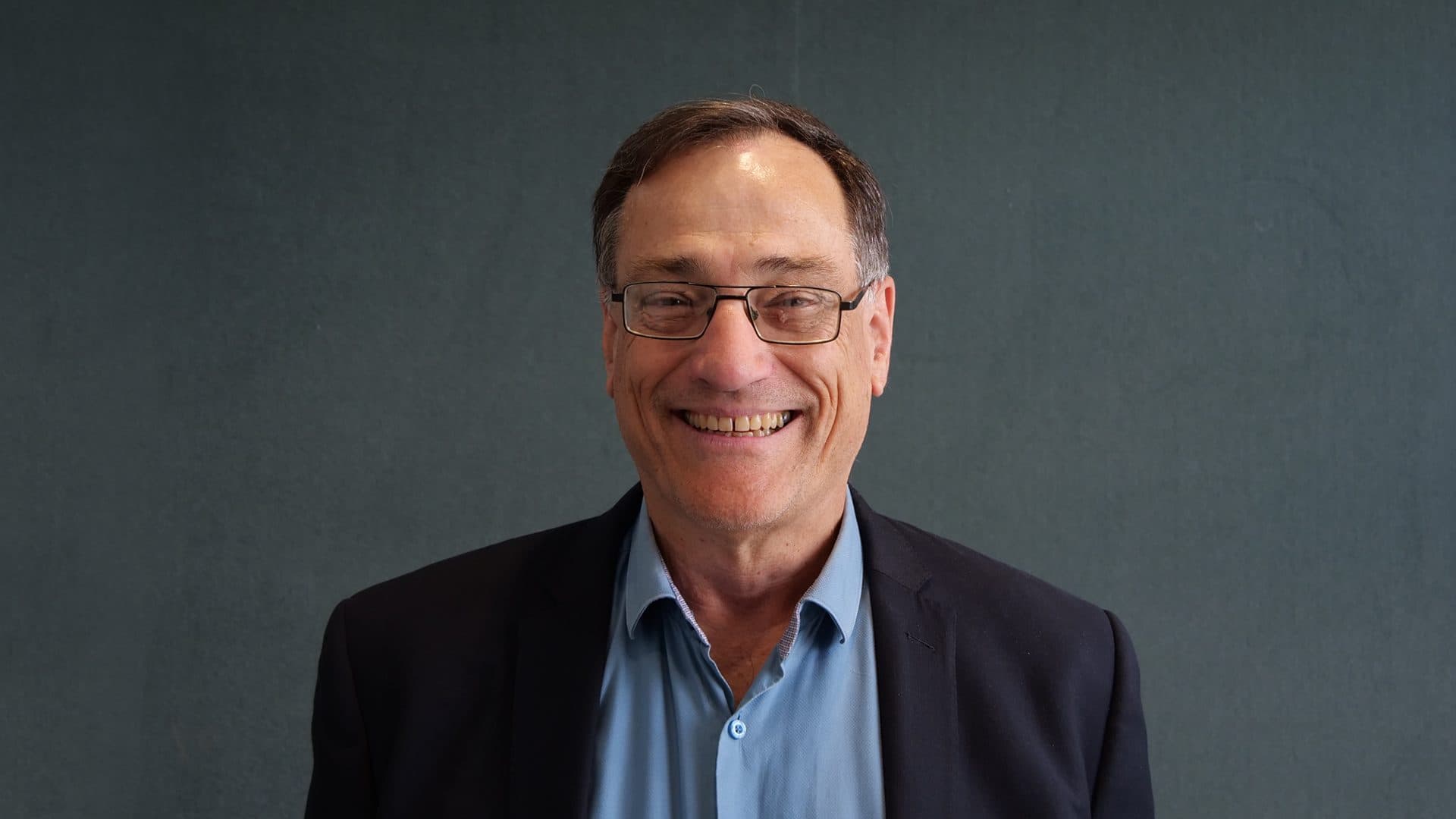 A man with brown hair wearing glasses, a blue shirt and navy jacket smiling for the camera with a dark grey internal background.