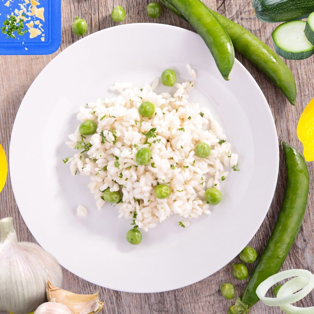 Rice, peas, parsley, and other vegetables on a white plate on a wooden table, surrounded by pea pods, courgettes, lemons, onions, and a chopping board with pieces of cheese and parley on it.