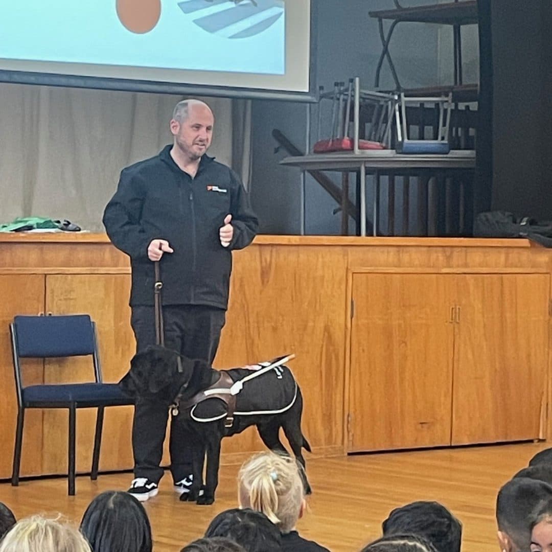 Dan Shepherd, wearing a black Blind Low Vision NZ branded jacket talks to schools children in a school hall with his guide dog Ezra, a black lab at his feet, wearing a Blind Low Vision NZ branded harness.