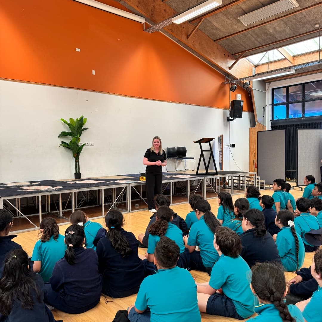 A woman stands on a stage in a school hall, presenting to a large group of students wearing teal uniforms who are seated on the wooden floor. A yellow guide dog harness rests on the stage floor near the speaker.