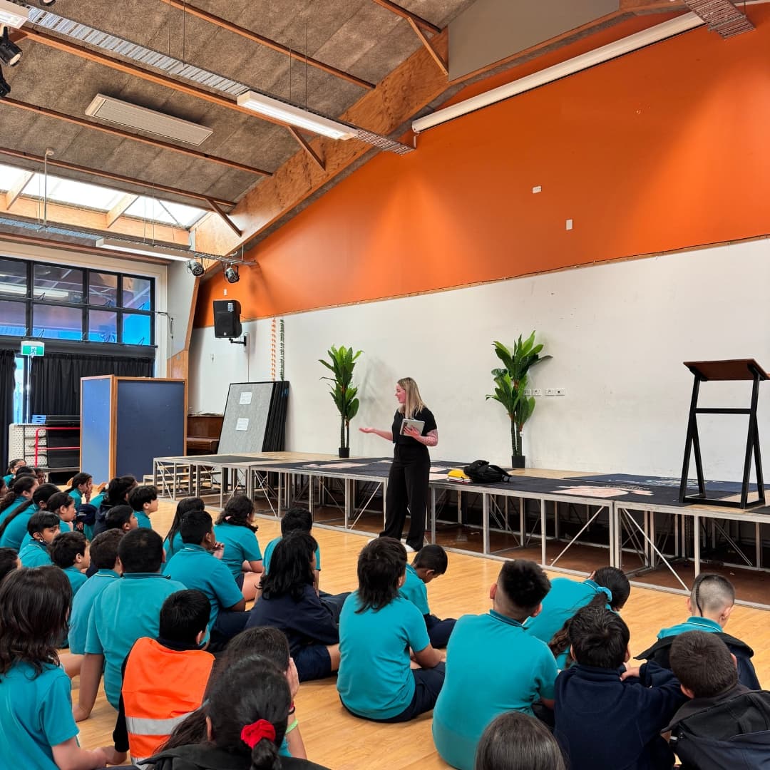 A presenter delivers a talk from a stage to a school assembly, holding a book in one hand and engaging with the students seated below.