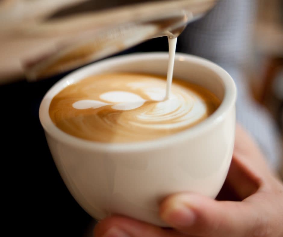 A hand holding a white cup as milk is poured into some coffee.