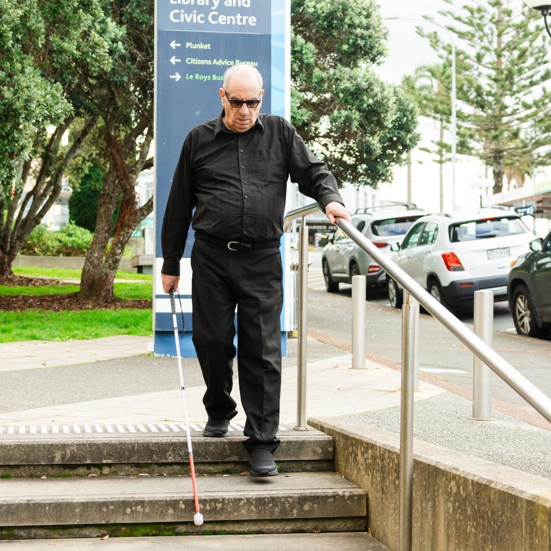 A man with a white cane walking down some concrete stairs in a suburban setting.
