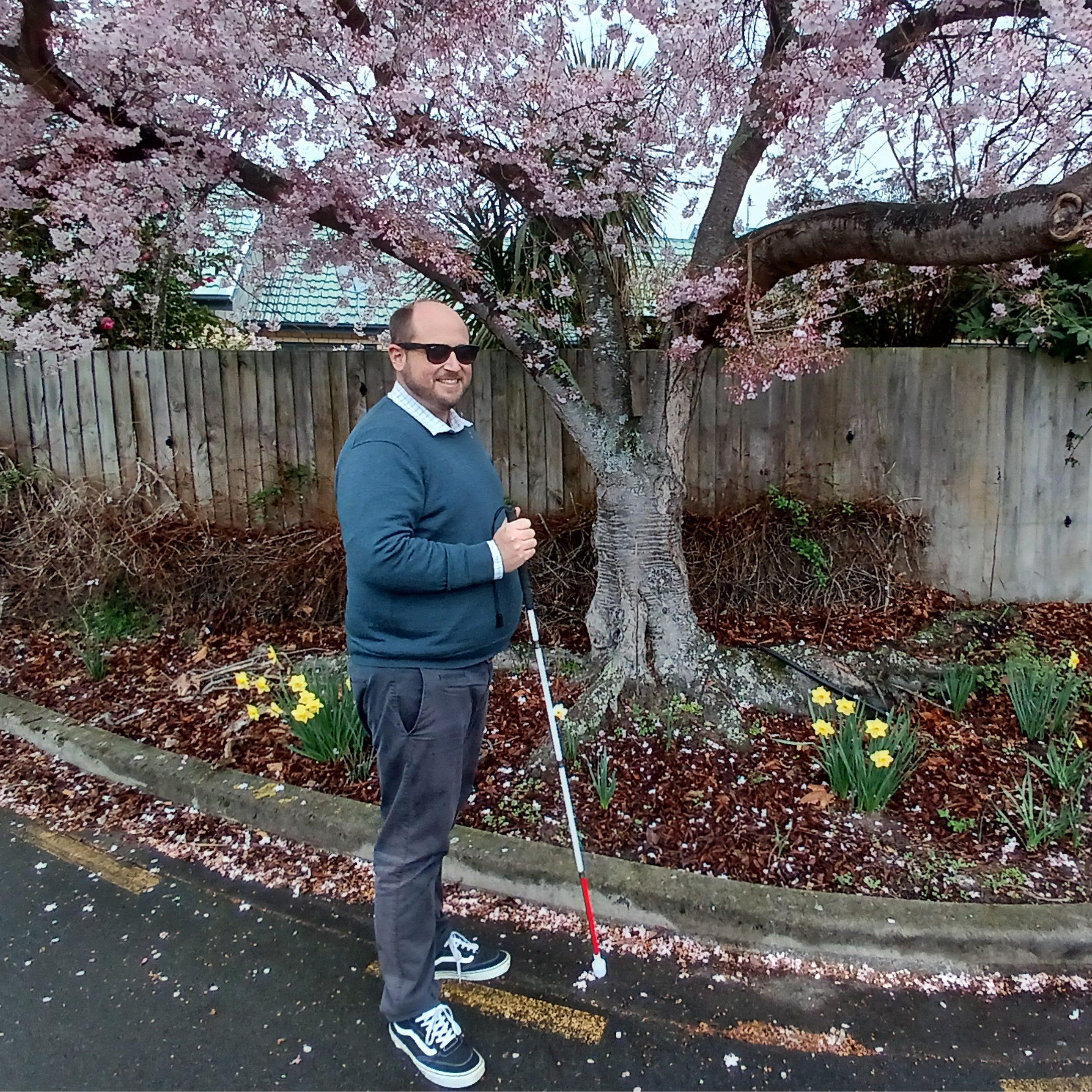 A man stands beneath a tree with cherry blossoms wearing dark glasses and holding a white cane. He is wearing a blue jumper and grey trousers.