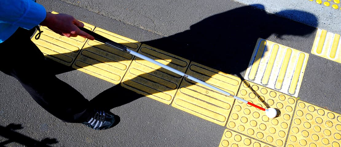 A client walks near the edge of a road and uses their white cane to help them navigate the tactile markings