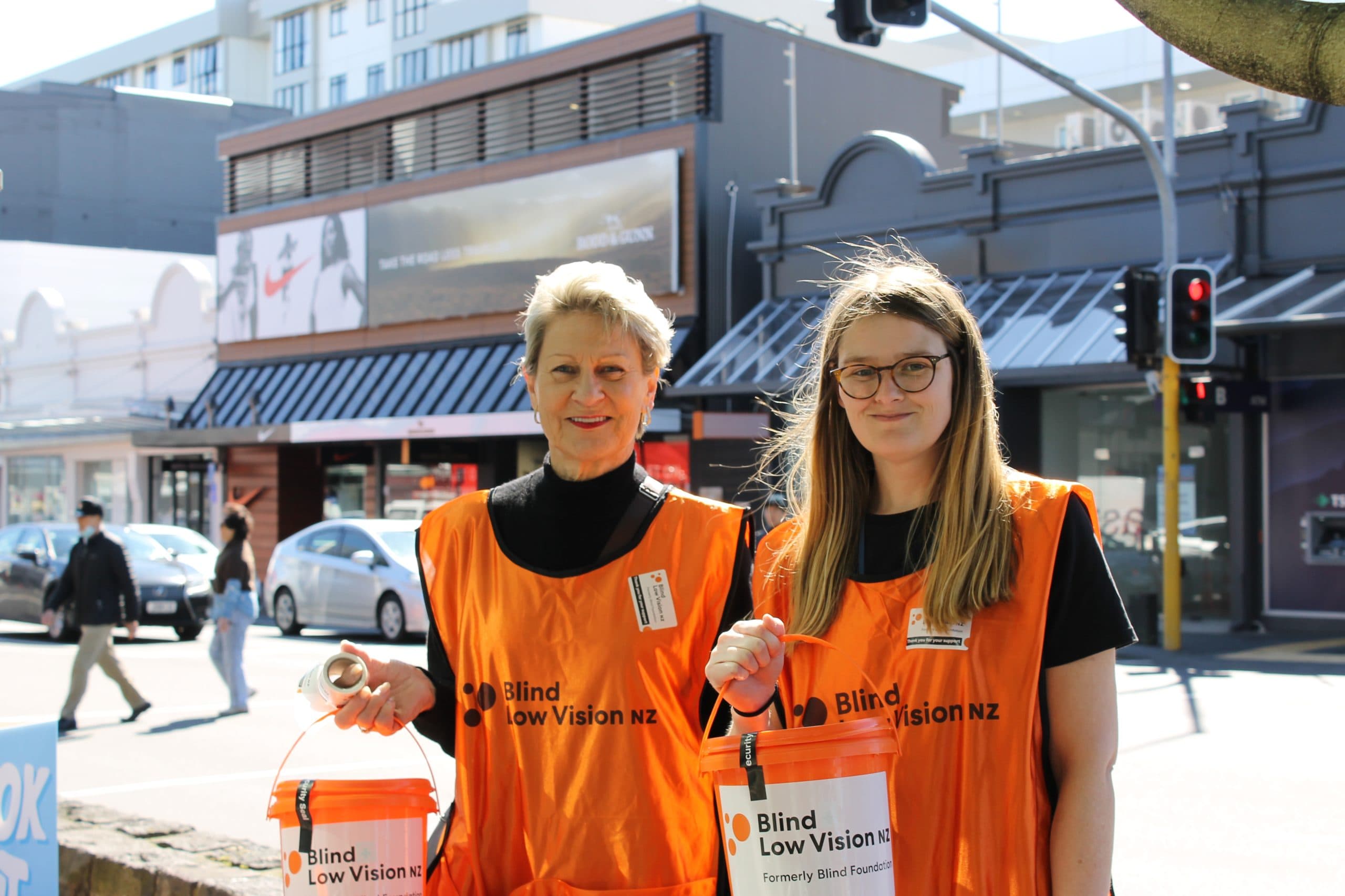 Two fundraising voluntees, Lynne and Hannah are collecting in Newmarket, they hold orange buckets and wear Blind Low Vision NZ bibs