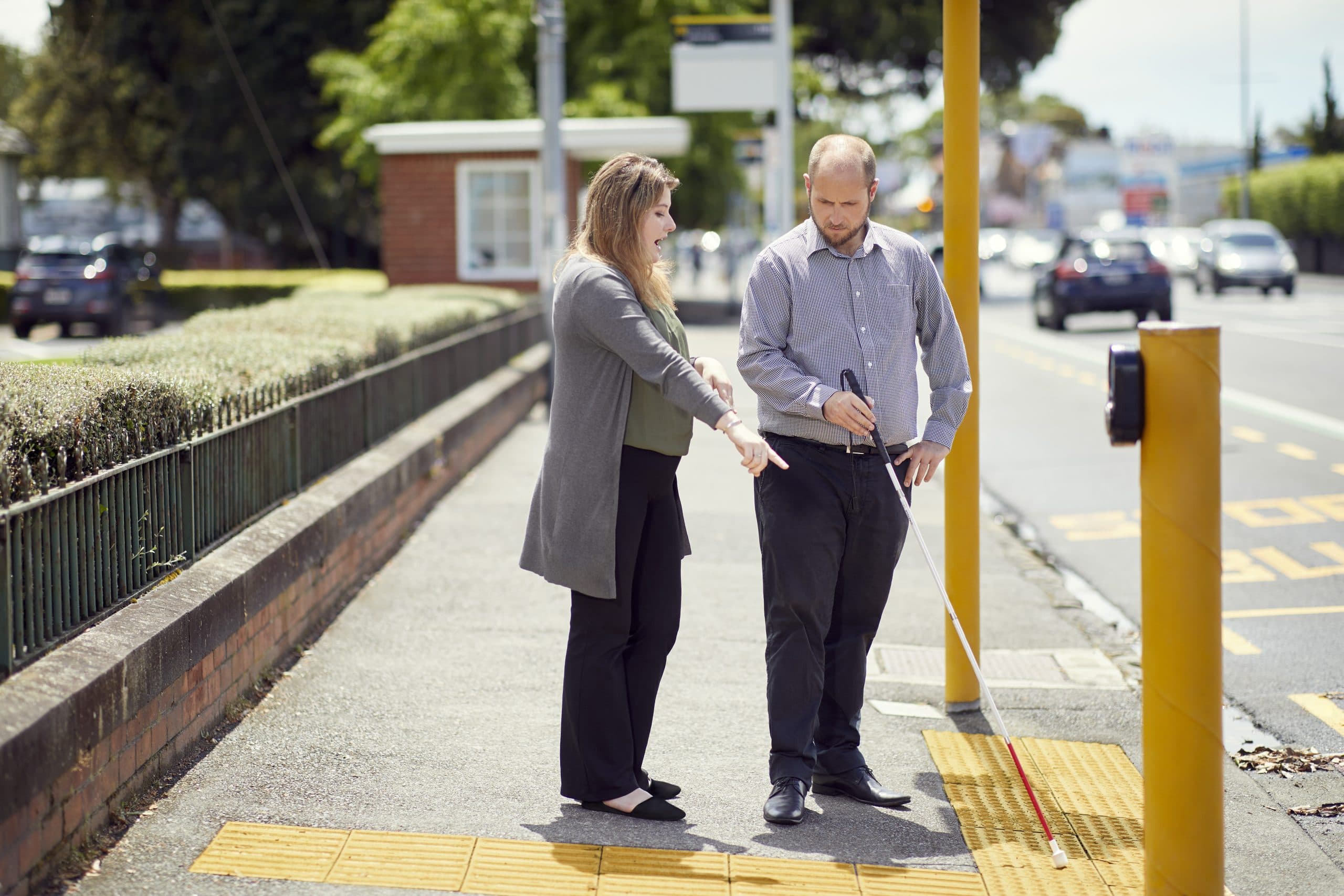 A male client is standing at a pedestrian crossing with his white cane and being taught how to use it by a female Blind Low Vision NZ staff member