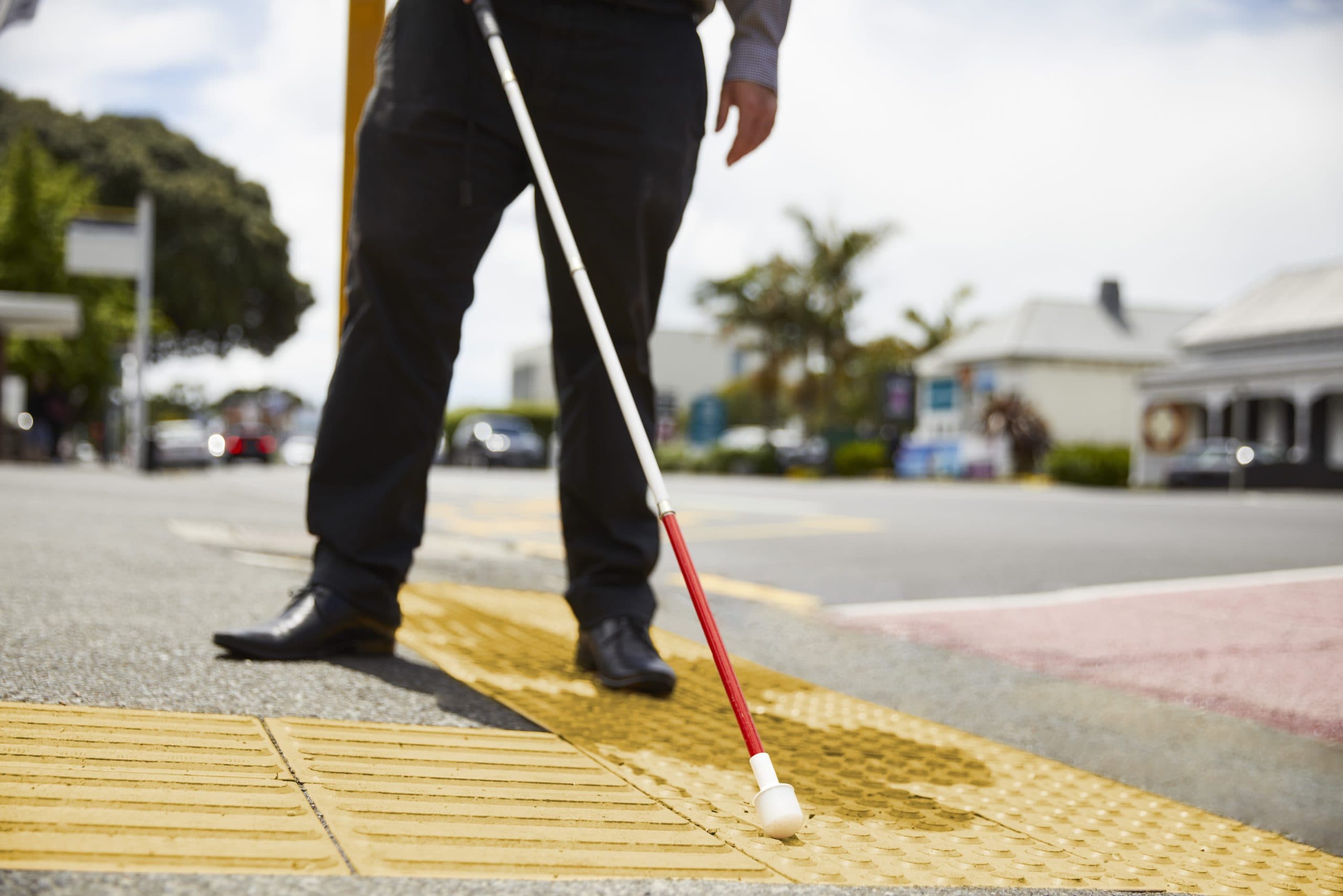 A male client is walking towards the camera on the pavement and his white cane is over the tactile markings