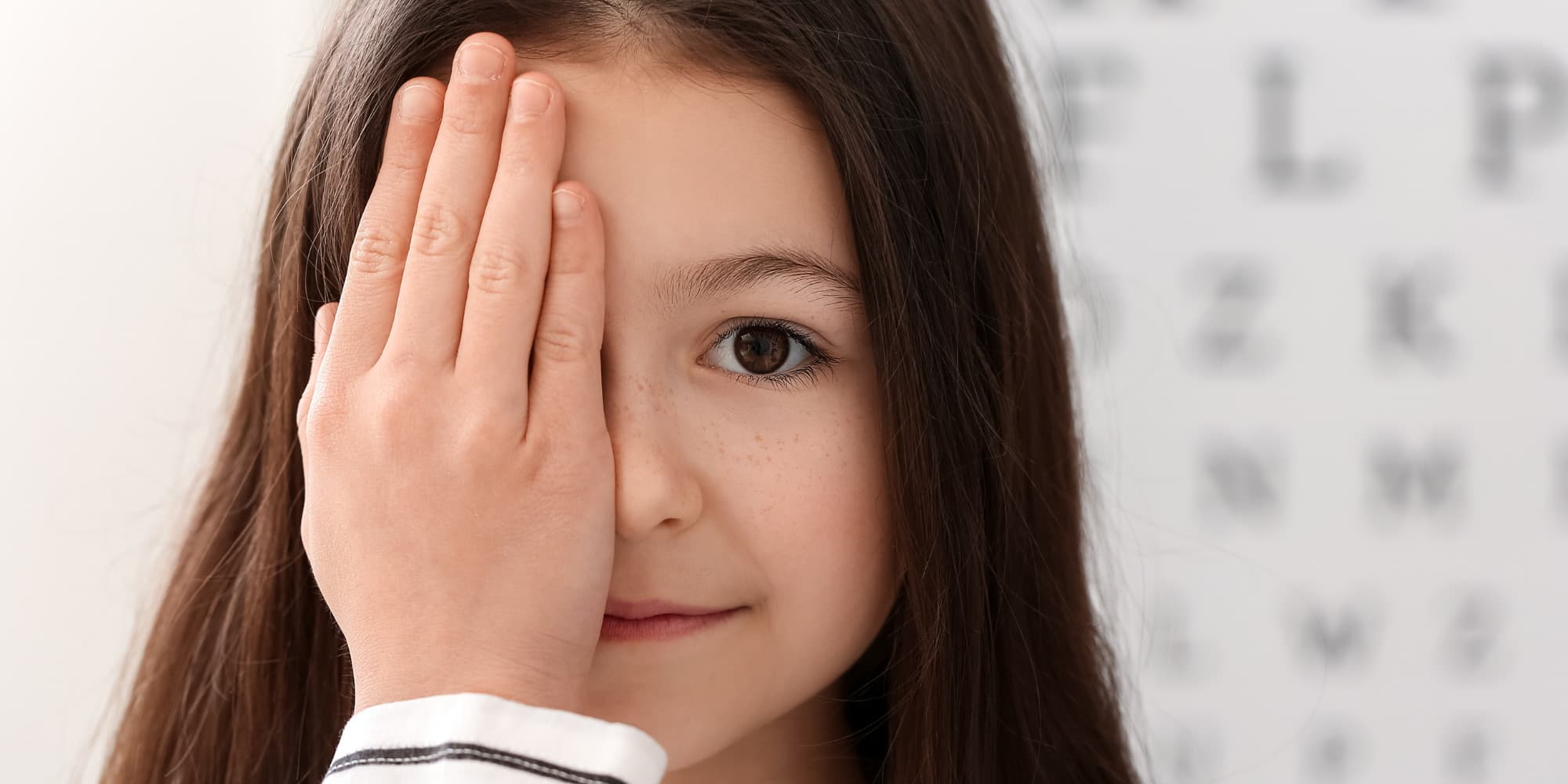 A young girl is looking towards the camera with one hand covering her right eye as if she is doing an eye test