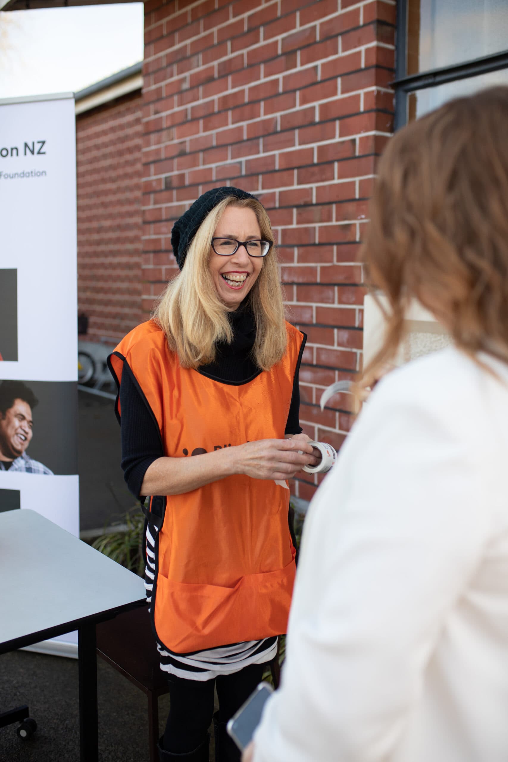A fundraising volunteer is standing at a table in the community, smiling towards someone who has come to talk about Blind Low Vision NZ's appeal
