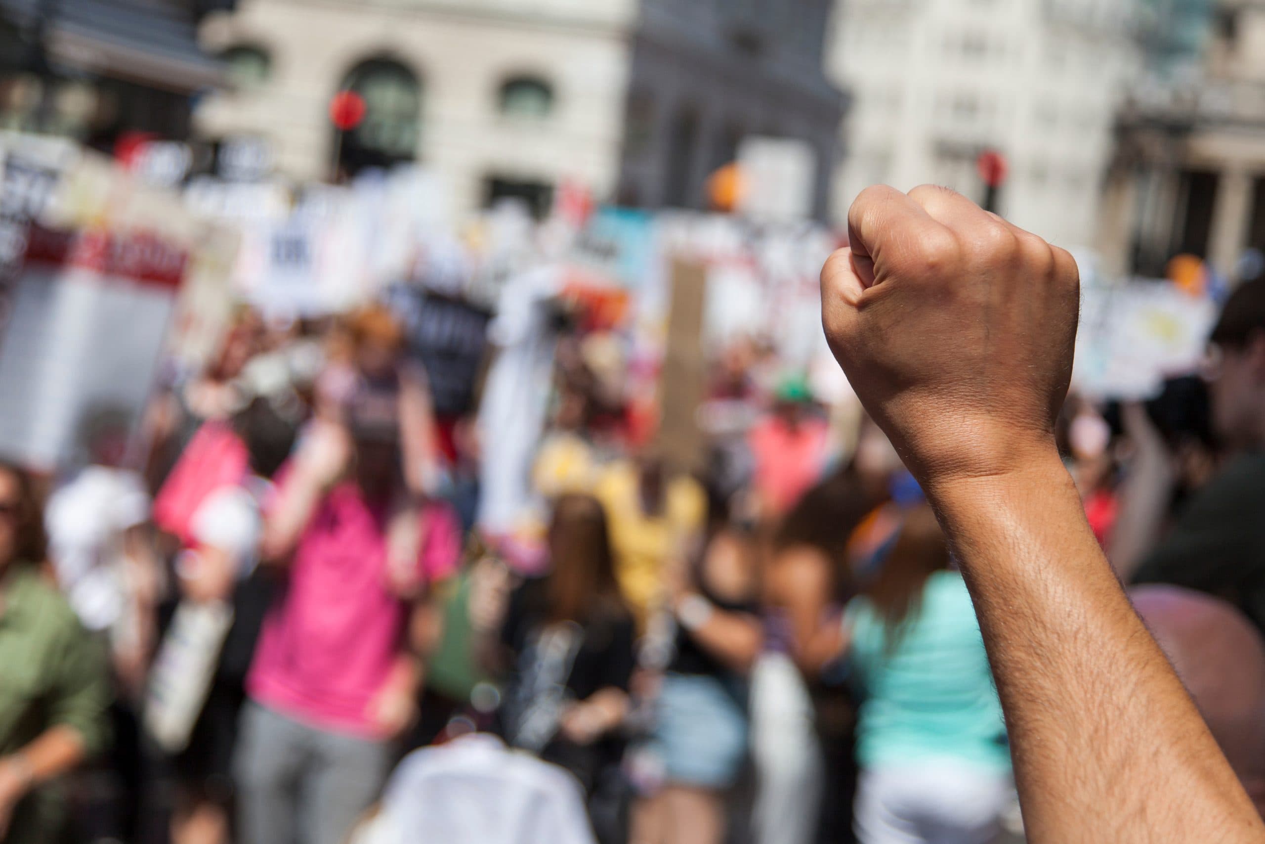 A raised fist in protest is in focus, the background shows a group of others campaigning