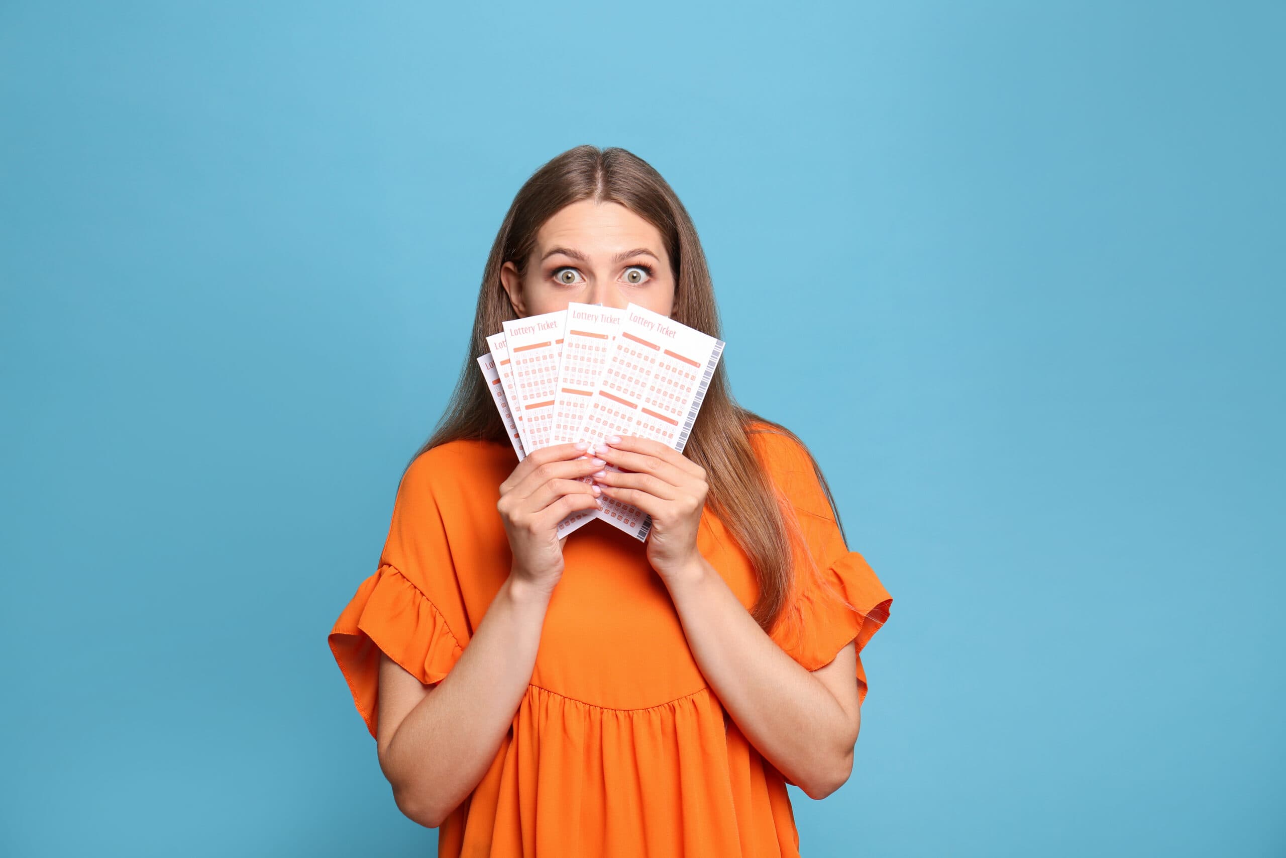 A young woman wearing a bright orange top holds lottery tickets and looks excitedly towards camera against a blue backdrop.