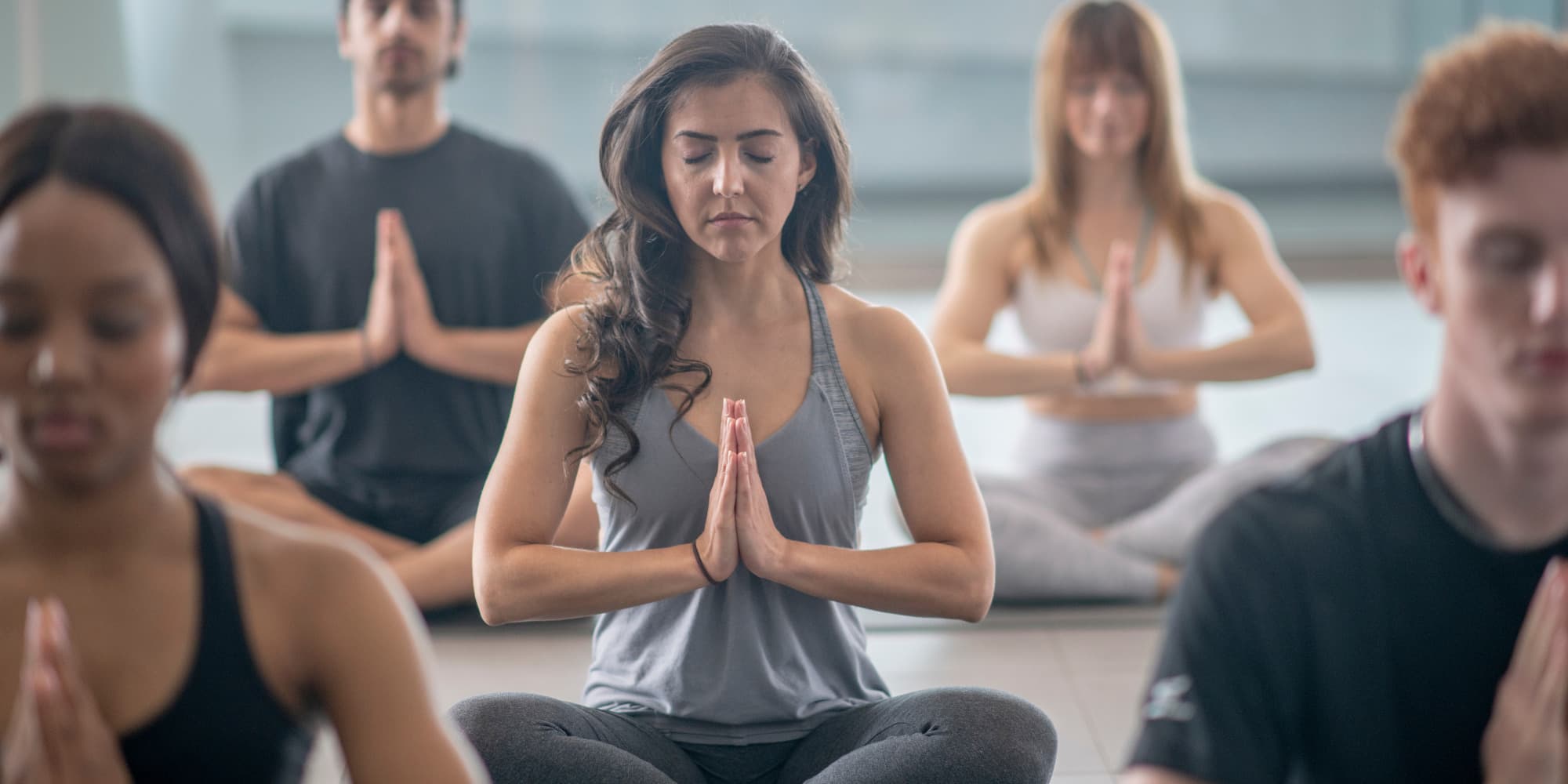 Several men and women sitting cross-legged with their hands together in a prayer pose with their eyes closed, meditating.