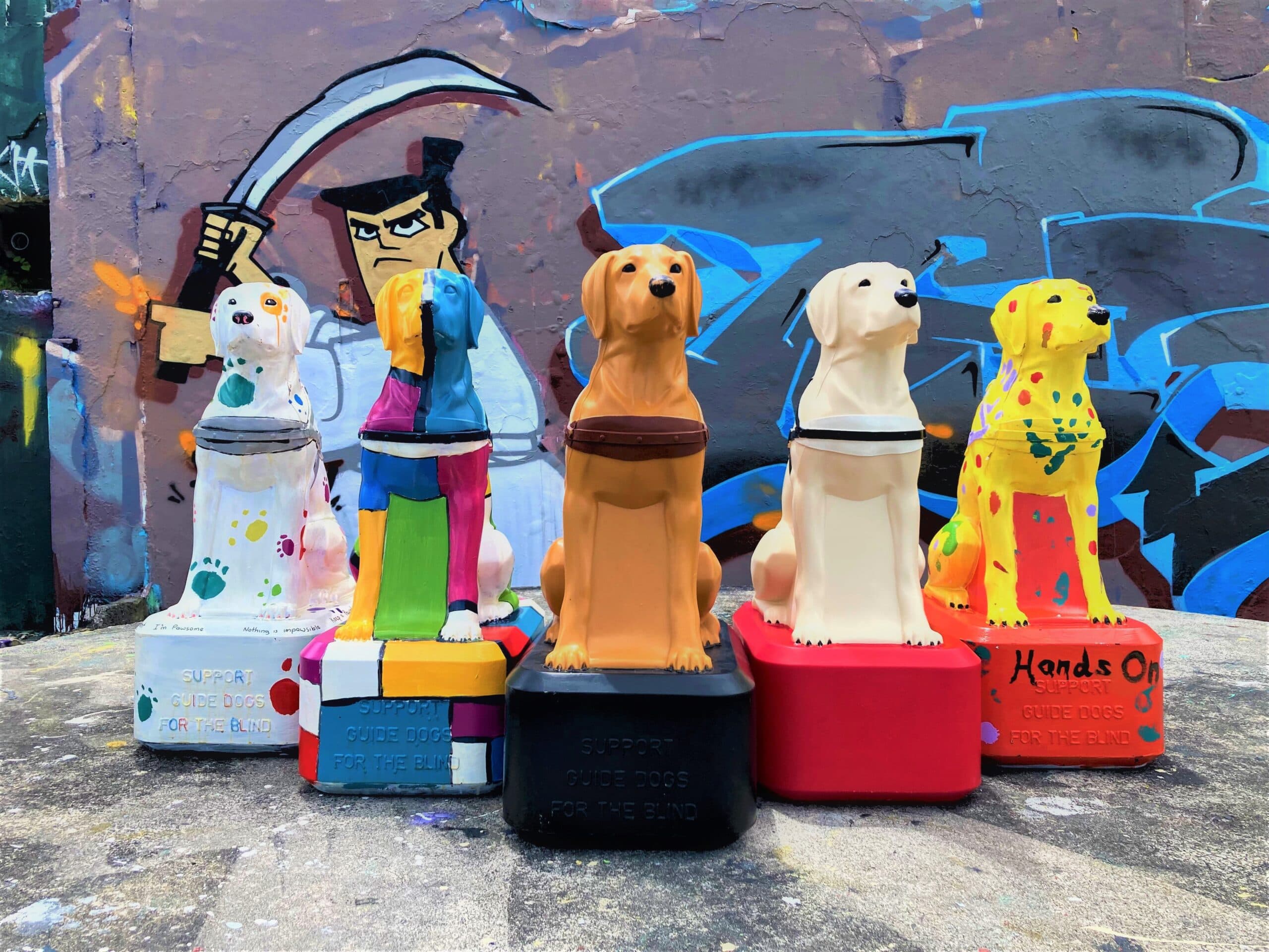 A group of life-sized dog-shaped collection boxes, Trudy Dogs, painted in bright colours, placed in front of an urban mural.