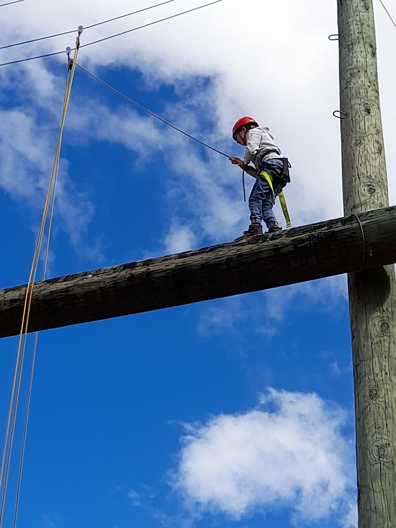 A young member walking along the high ropes beam approximately ten meters above ground, supported by a harness and ropes, with blue sky and white clouds in the background.