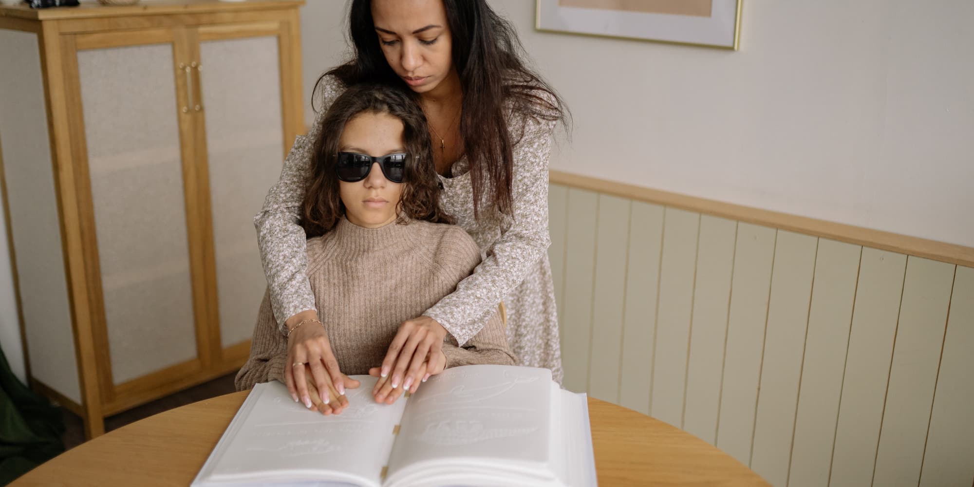 A sighted mother guiding her blind daughter's hands over a braille book.