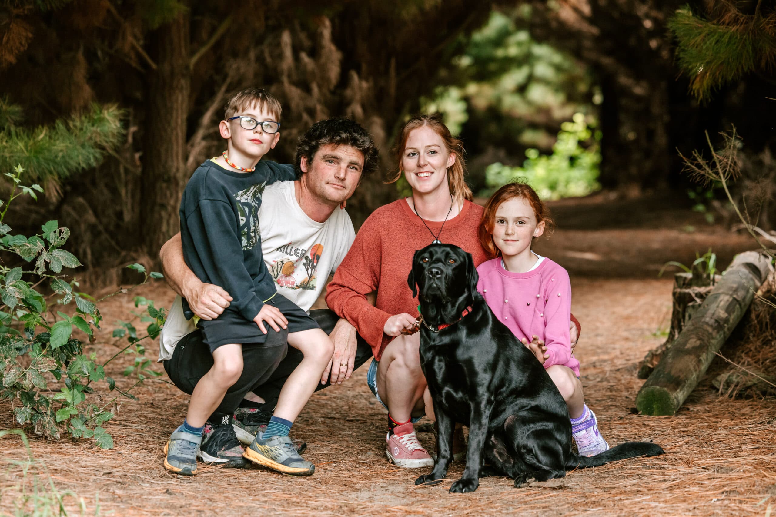 A young family with their two children and black Labrador guide dog, posing for the camera while out for a trek in the woods.