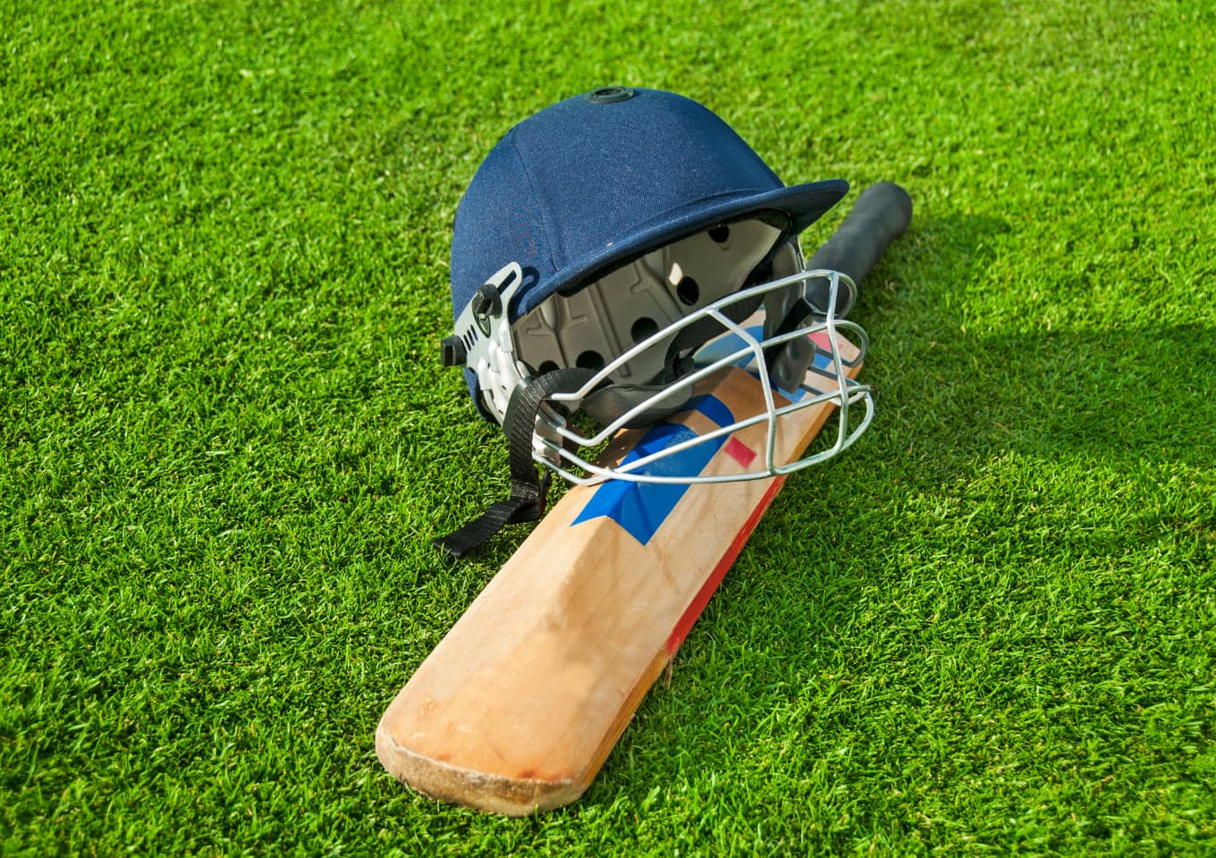 Cricket helmet resting on a cricket bat placed on bright green cricket pitch.