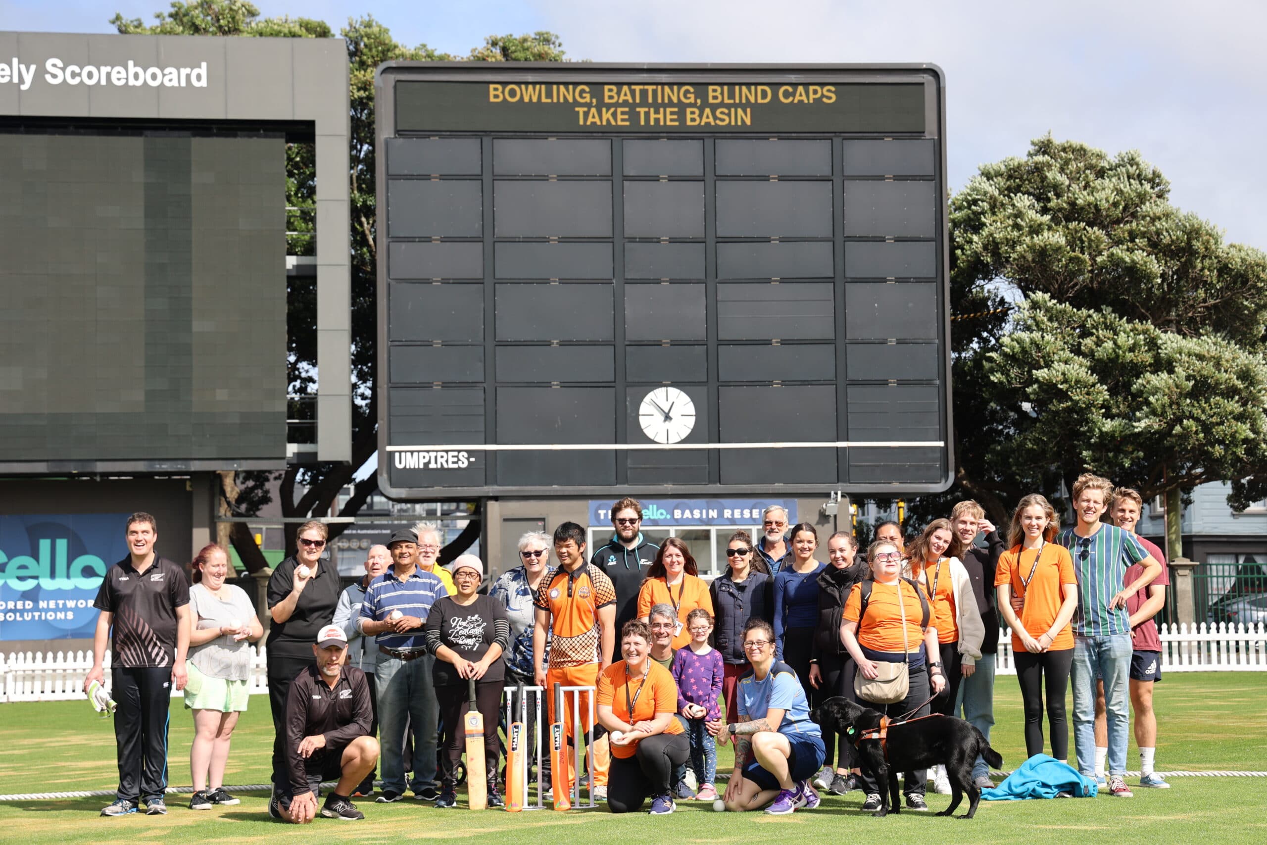 A group of Blind Low Vision NZ members, friends, family and staff standing in front of the Basin Reserve scoreboard that reads “ Bowling, Batting, Blind Caps Take the Basin”. It's a sunny day even though a few clouds appear in the background.