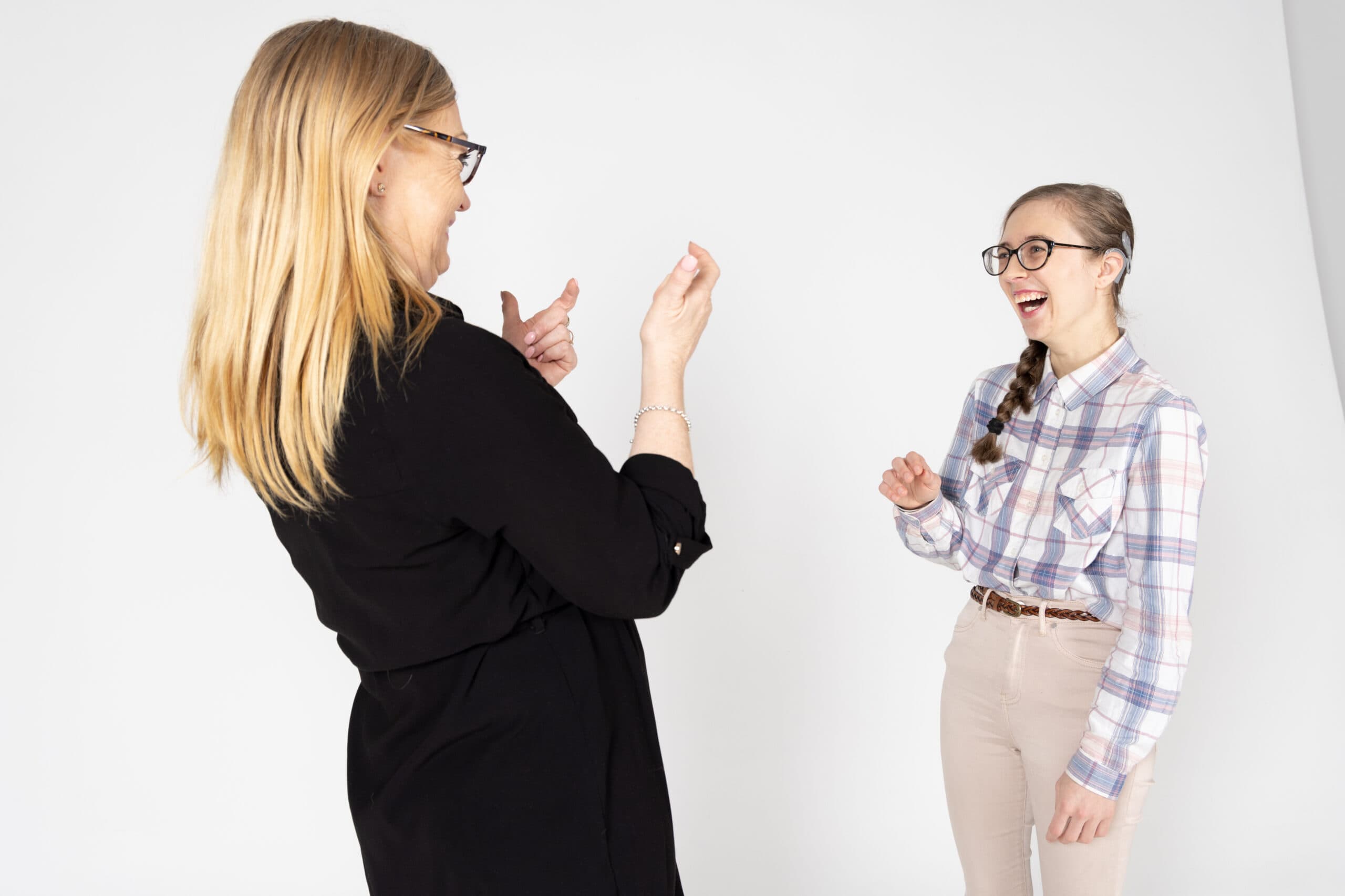 A young woman wearing glasses and a checked shirt having a laugh with another woman in black clothes as they use sign language.