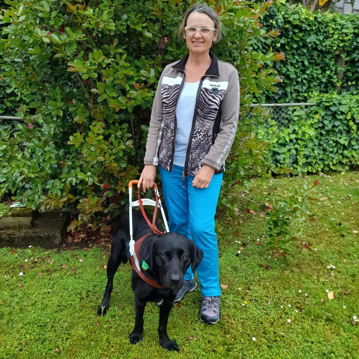 Our client, Rhonda Comins standing outdoors with a tree in the background, accompanied by her guide dog, a black Labrador in a harness. Rhonda is smiling for the camera.