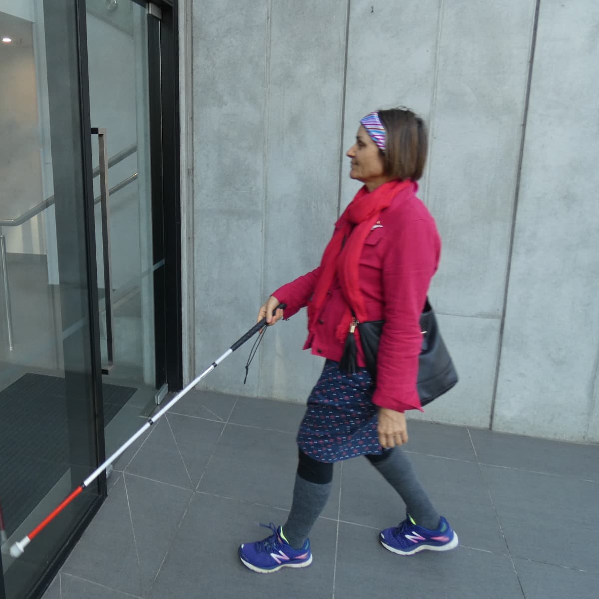 Rhonda, wearing a bright pink jacket and holding her white cane at the entrance to a building, locating the door.
