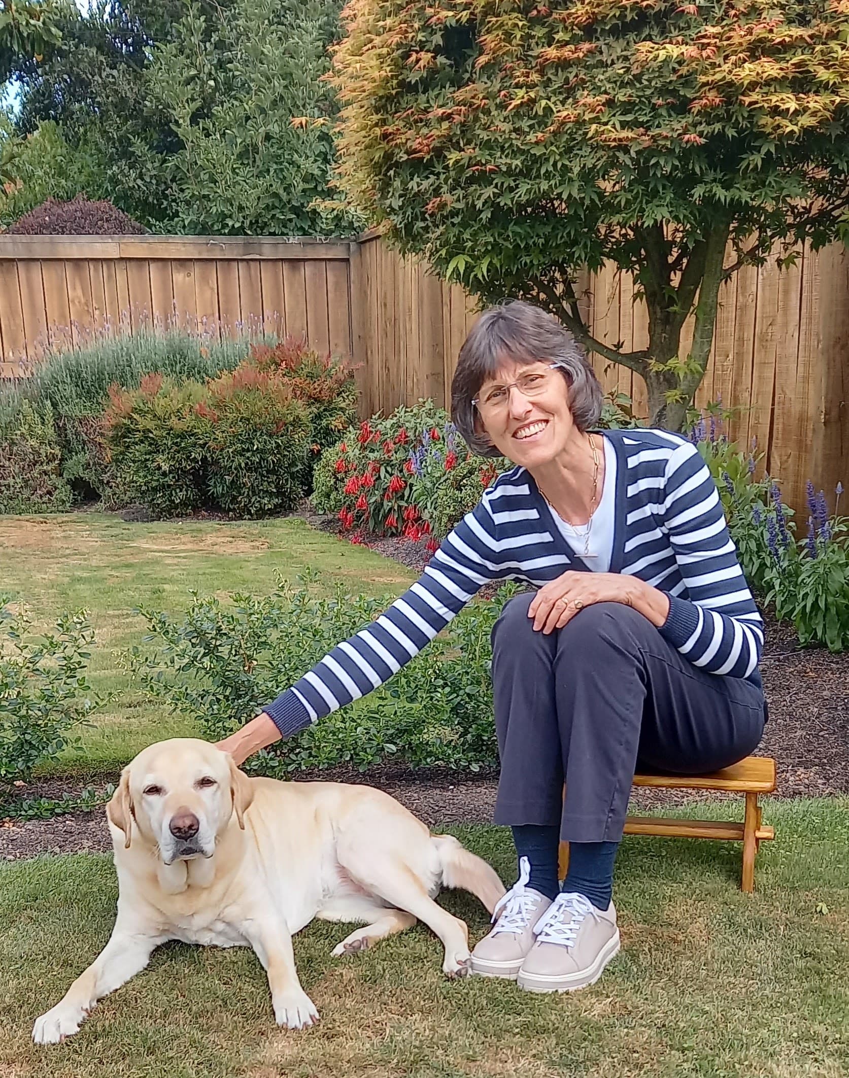 Visionary Ruth sitting in her garden, smiling for the camera. She is patting a golden retriever who is lying at her feet.
