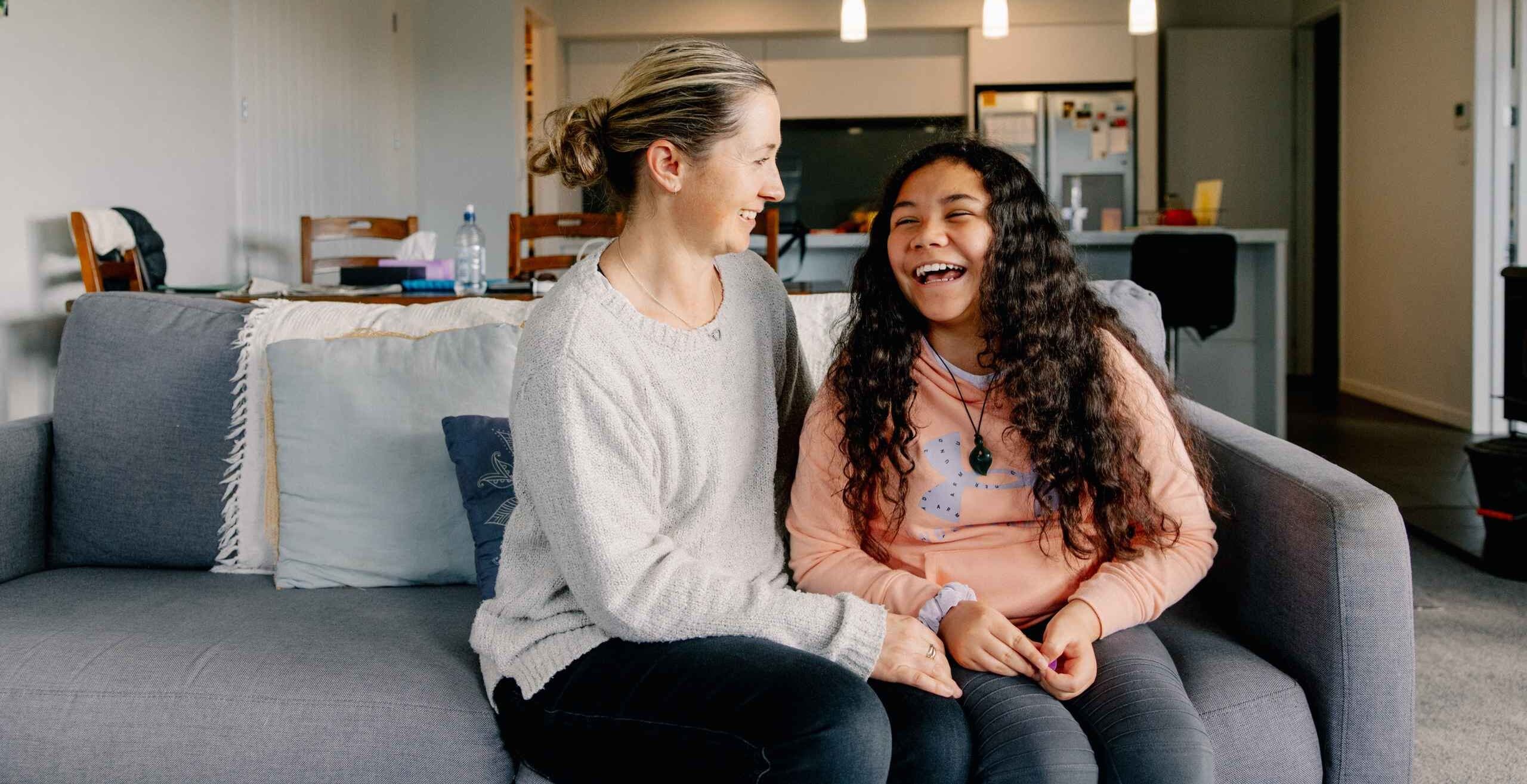 Marama and her mum sitting on a couch at home, laughing.