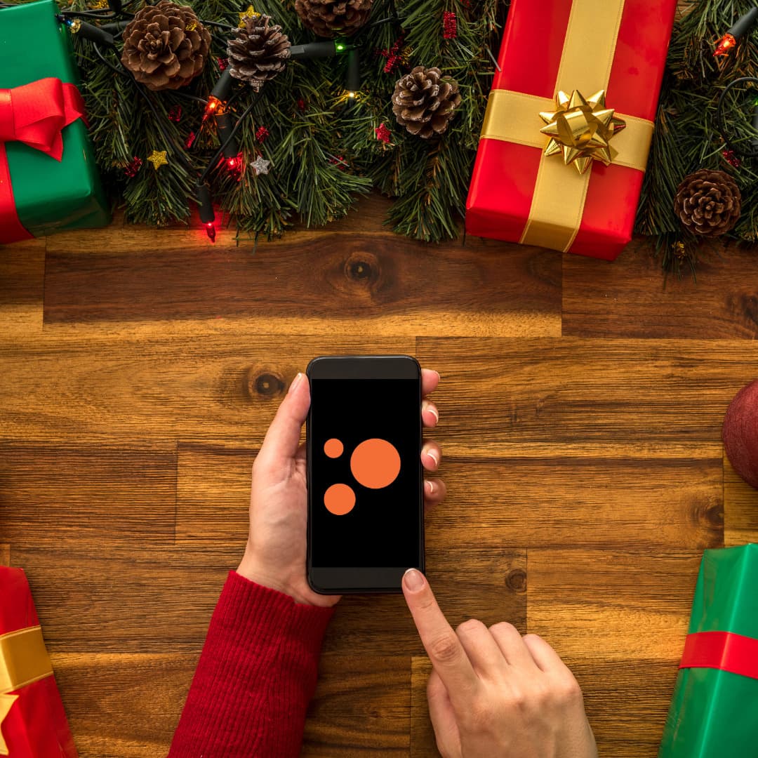 An image of a hand holding a smartphone with the Blind Low Vision NZ logo on the screen, surrounded by giftwrapped Christmas presents.