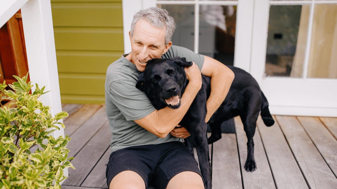 Peter Jones sitting on his porch with his guide dog Ayla, a black labrador. Peter has his arms around Ayla.