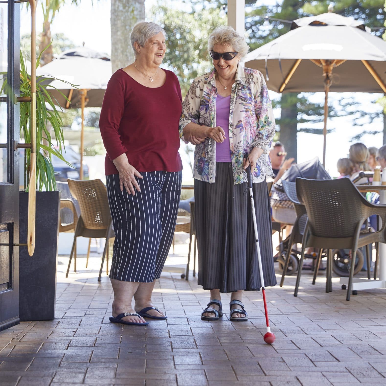 Two women walking through an outdoor cafe setting. One is using a white cane.