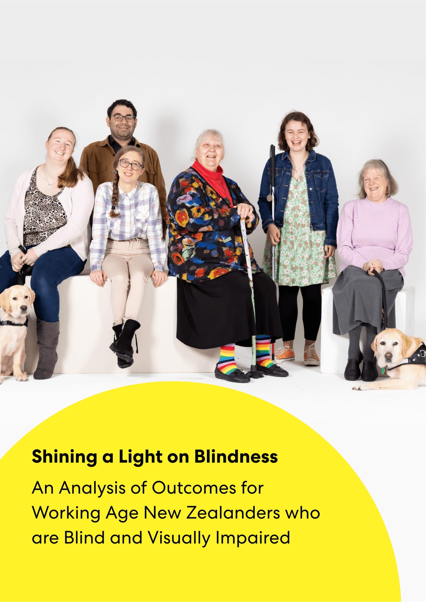 A diverse group of people sitting together, smiling for the camera. Two have dogs at their feet, while another has a walking cane in her hands, and another has a white cane.