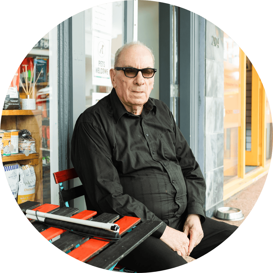 An older gentleman sitting at a red and black table outside a shop. His white cane is on the table.