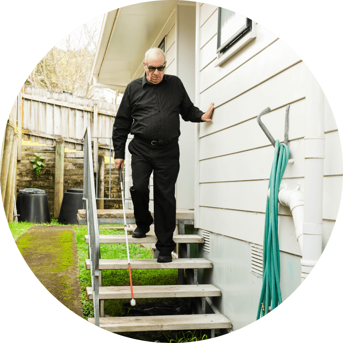 A man dressed in black walking down the stairs outside of his home with his white cane.